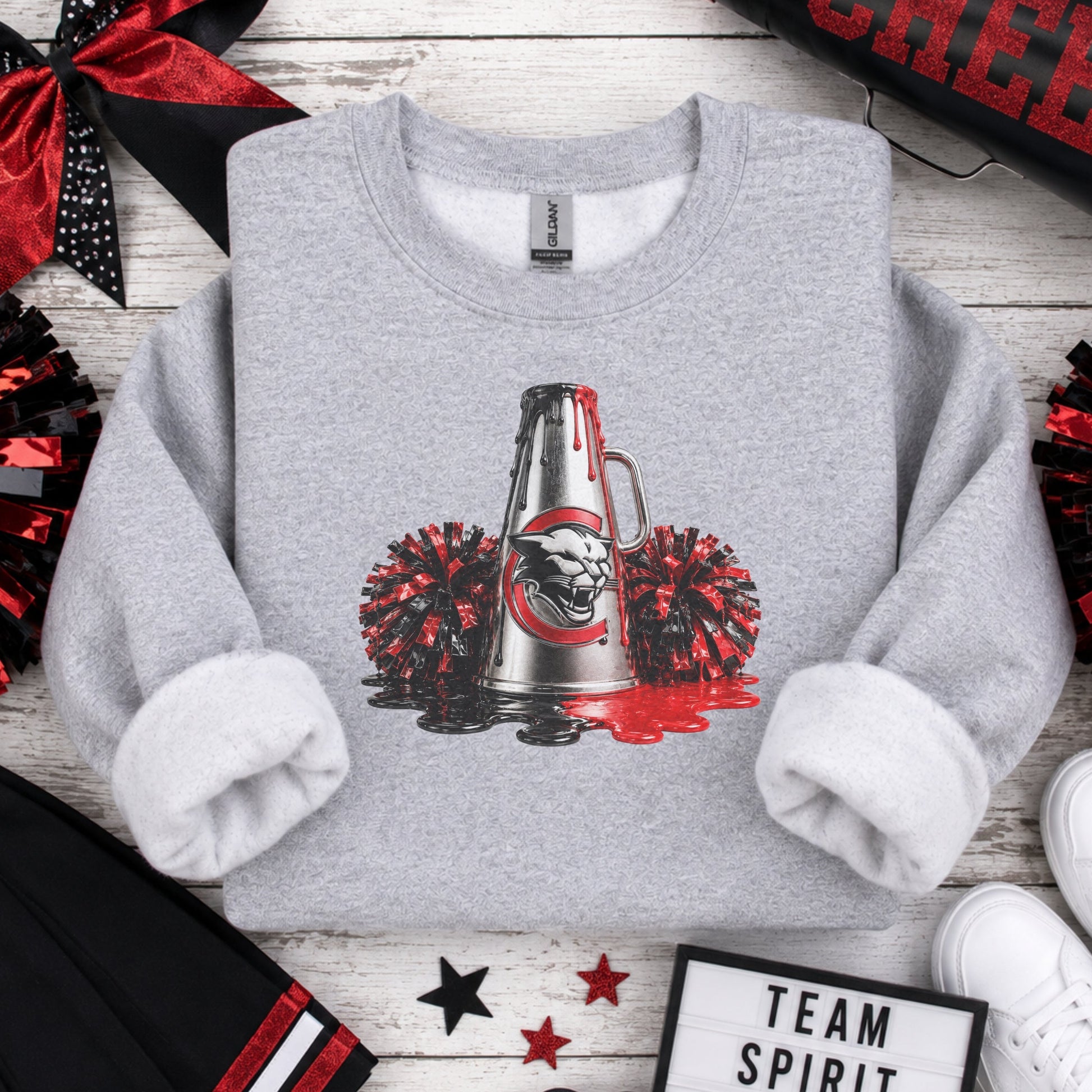 A heather gray crewneck sweatshirt with rolled cuffs, featuring a graphic of a silver cheerleader megaphone with red paint splatter and a black red and white Charleroi Cougar logo, surrounded by red and black pom-poms. The sweatshirt is styled in a flat-lay on a white wood background with cheerleader accessories, white sneakers, and a 'Team Spirit Win' sign.
