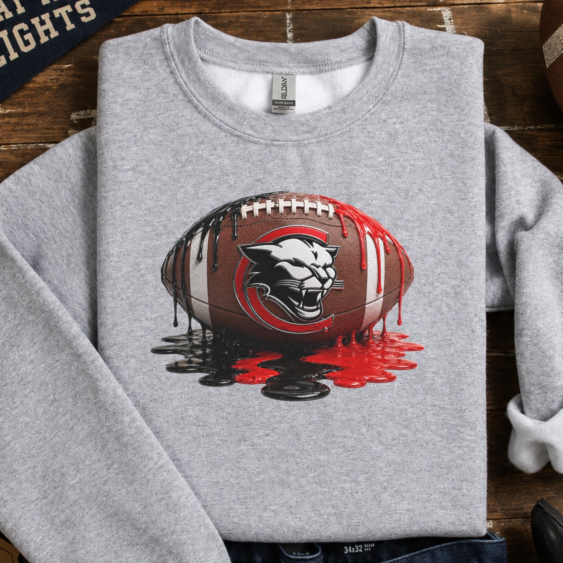 A heather gray crewneck sweatshirt featuring a graphic of a football with the Charleroi cougar logo and red paint splatter, styled in a flat lay on a wooden background with jeans, a football, tickets, and a pennant.