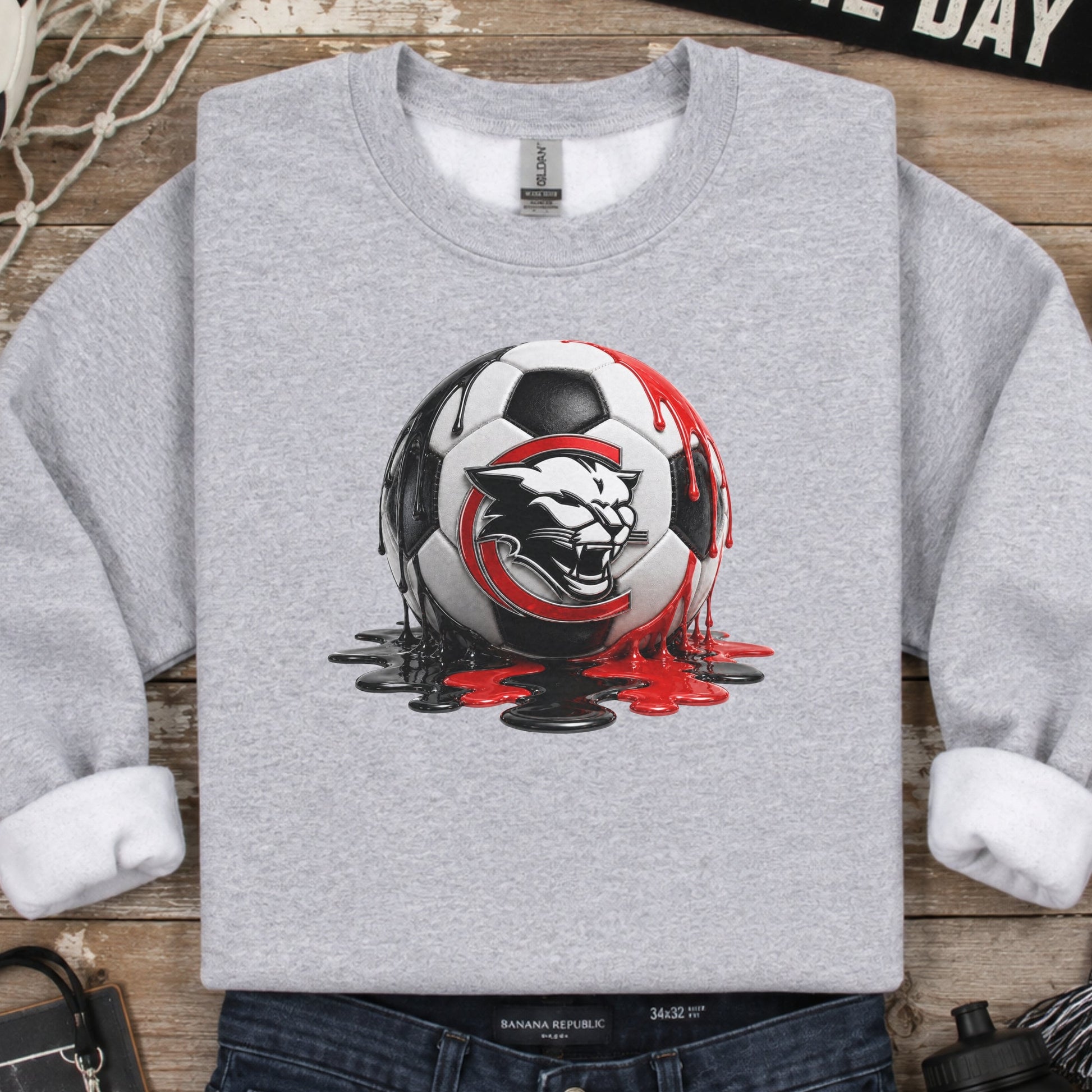 A heather gray crewneck sweatshirt with a graphic of a soccer ball and a wild cat head, styled in a flat lay with jeans, a scarf, a water bottle, and other soccer-themed props on a rustic wooden background.