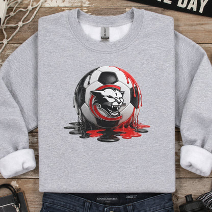 A heather gray crewneck sweatshirt with a graphic of a soccer ball and a wild cat head, styled in a flat lay with jeans, a scarf, a water bottle, and other soccer-themed props on a rustic wooden background.
