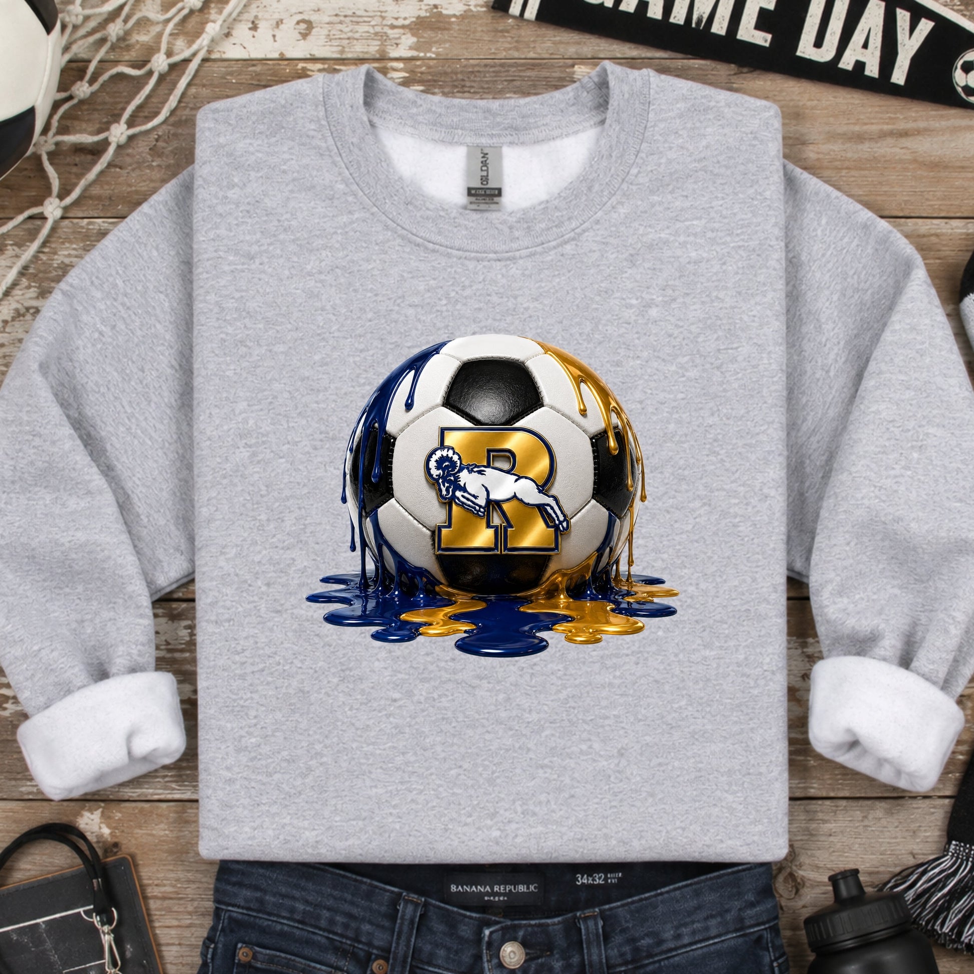 A heather gray crewneck sweatshirt featuring a graphic of a soccer ball with the Ringgold Rams R Ram Logo and blue/gold paint drips, styled in a flat lay with jeans, a scarf, a soccer ball, and other sports-themed props on a wooden background.