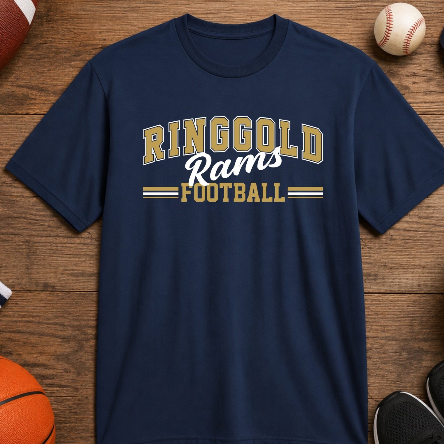 A navy blue short-sleeve t-shirt with 'RINGGOLD Rams FOOTBALL' printed in vegas gold and white on the chest, displayed in a flat-lay arrangement on a wooden surface surrounded by sports items like a basketball, soccer ball, baseball, whistle, and shoes.