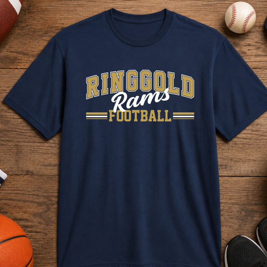 A navy blue short-sleeve t-shirt with 'RINGGOLD Rams FOOTBALL' printed in vegas gold and white on the chest, displayed in a flat-lay arrangement on a wooden surface surrounded by sports items like a basketball, soccer ball, baseball, whistle, and shoes.