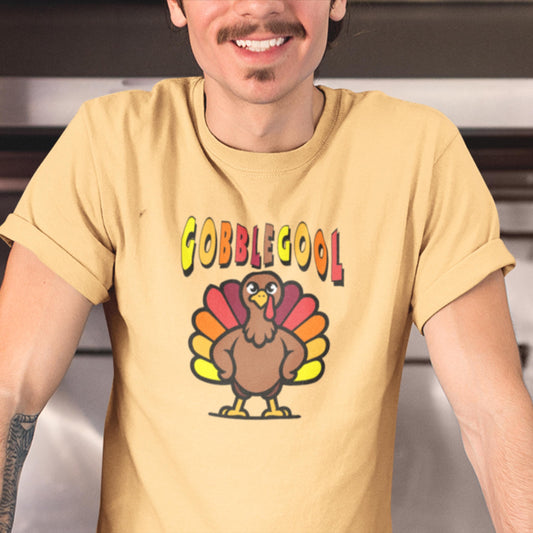 Tan-colored T-shirt featuring a humorous cartoon turkey with the word &#39;GOBBLEGOOL&#39; in bold, colorful letters above it. The turkey has a confident pose with wings on its hips. Perfect for a fun, Italian-themed Thanksgiving look.