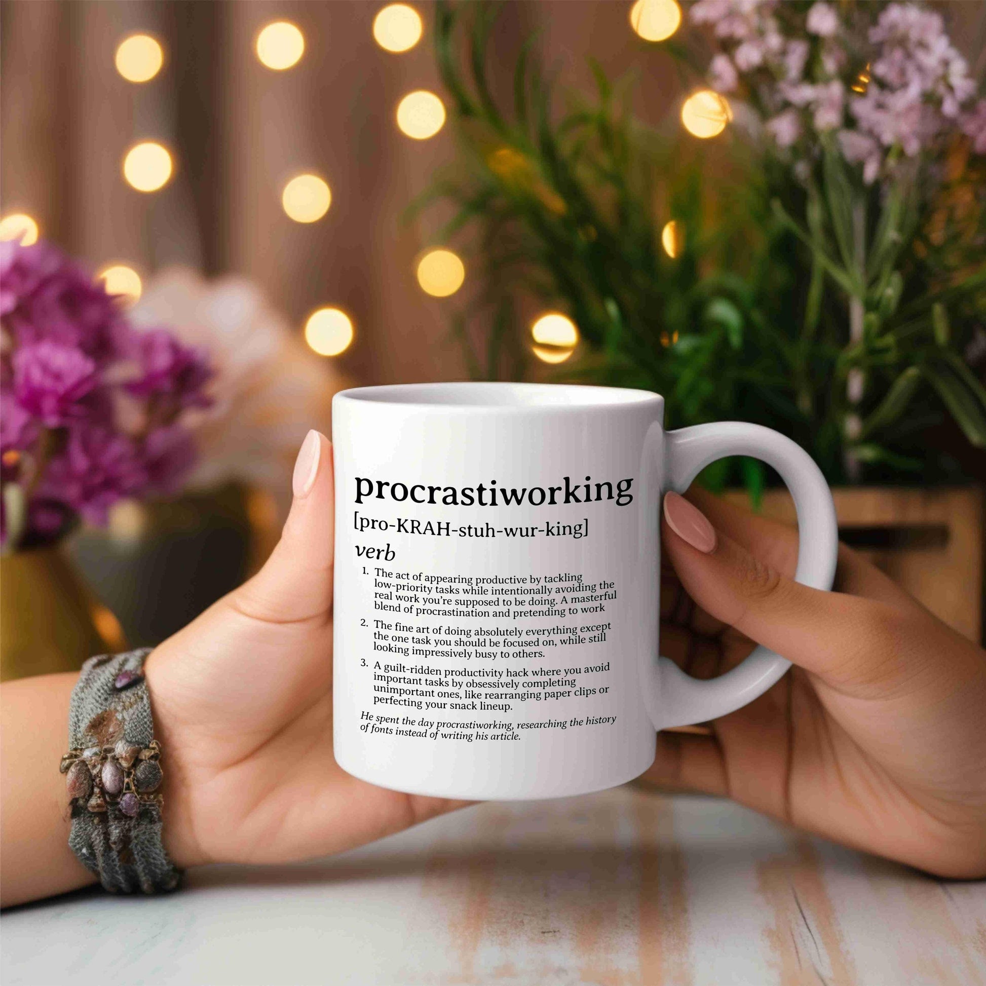 White ceramic mug with a humorous &#39;procrastiworking&#39; definition, held against a blurred floral background with bokeh lights.