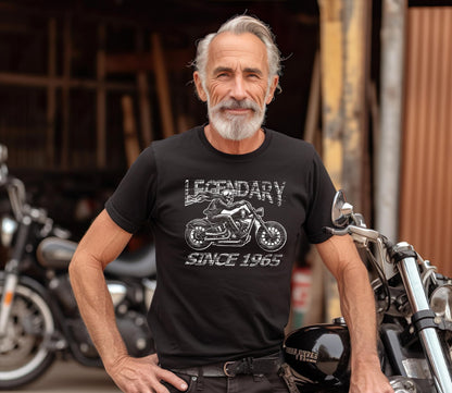 A older man with a  smile wearing a black T-shirt that reads Legendary Since 1965 with a graphic of a motorcycle and a distressed vintage design He is standing beside a classic motorcycle in a  garage setting, exuding timeless biker style and charm.