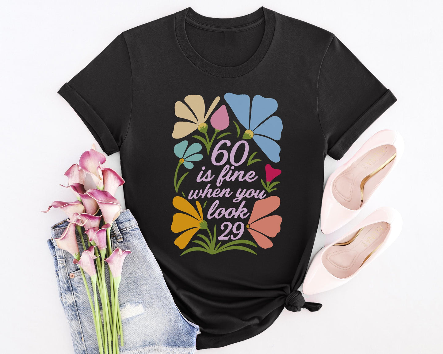 Black t-shirt with floral design and “60 is fine when you look 29” quote, styled with denim and pink heels.