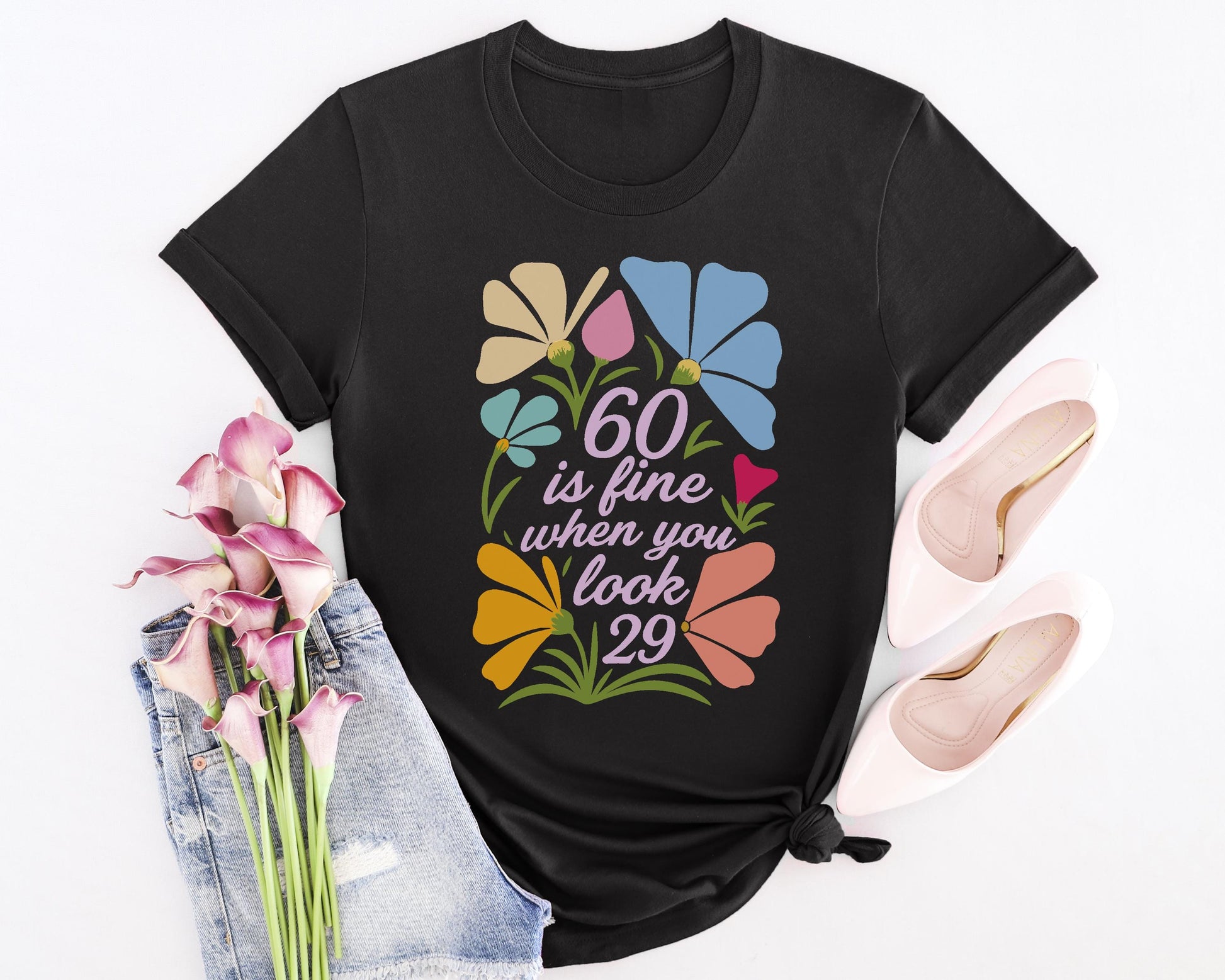 Black t-shirt with floral design and “60 is fine when you look 29” quote, styled with denim and pink heels.