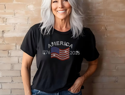 Woman wearing black rhinestone America 250th t-shirt with red, white, and blue flag, smiling against brick wall.