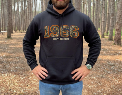 Man wearing black hunting hoodie with camouflage 1966 graphic and “Born to Hunt” text, outdoor forest setting, rugged hunting apparel.  Great gift for a hunter celebrating his/her's 60th birthday