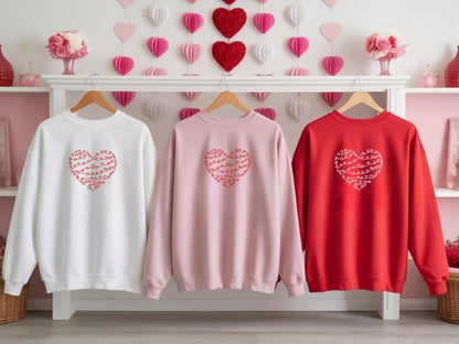 Three minimalist Valentine’s Day heart line sweatshirts in white, pink, and red hanging on a rack with heart decor background