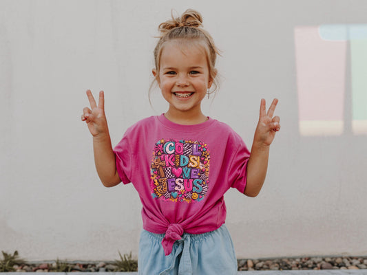 Child wearing a crunchberry t-shirt with colorful "Cool Kids Love Jesus" text in a ransom collage letters, leopard and checkered patterns