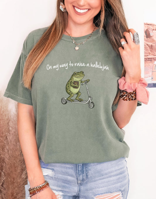 Woman wearing a t-shirt featuring a cute frog holding a Bible and riding a scooter with text On my way to raise a hallelujah.